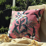 Honolulu Floral Polyester Cushion in pink, square outdoor cushion with tropical white blooms, dark leaves and palm fronds, shown outdoors against a woven throw and dark fence backdrop.