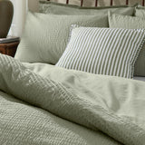 Hebden Cotton Plain Duvet Cover Set in bay green, showing the seersucker-textured cover and matching pillowcases on a bed with a striped accent cushion.