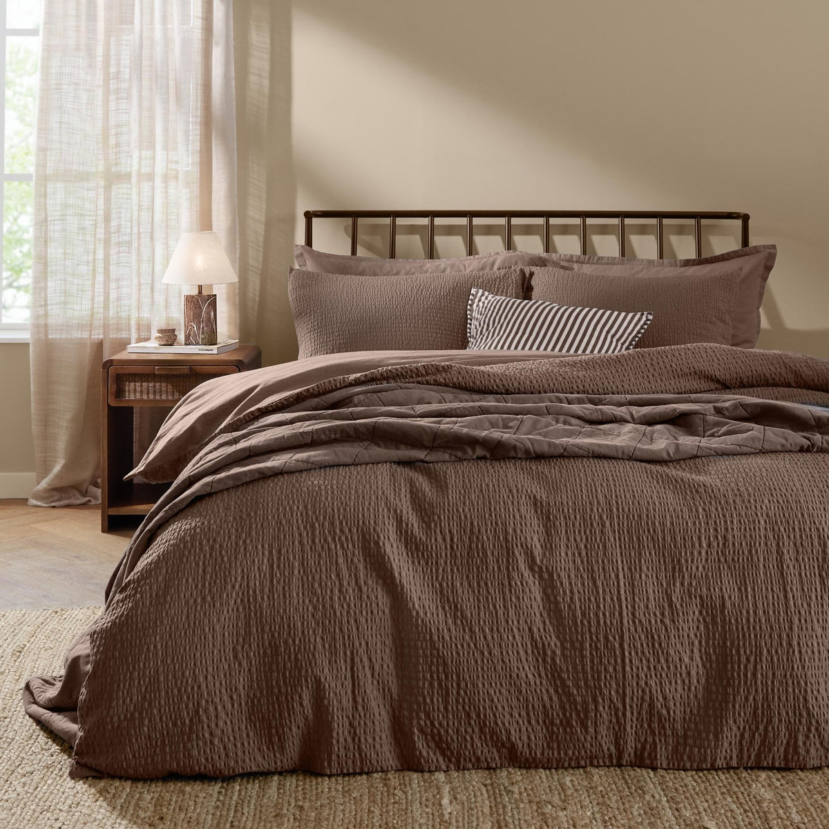Hebden Cotton Plain Duvet Cover Set in cocoa brown on a bed with matching pillowcases and a striped accent cushion, shown in a bedroom with a dark metal headboard.