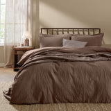 Hebden Cotton Plain Duvet Cover Set in cocoa brown on a bed with matching pillowcases and a striped accent cushion, shown in a bedroom with a dark metal headboard.