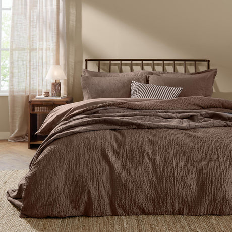 Hebden Cotton Plain Duvet Cover Set in cocoa brown on a bed with matching pillowcases and a striped accent cushion, shown in a bedroom with a dark metal headboard.