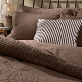 Hebden Cotton Plain Duvet Cover Set in cocoa brown, shown on a bed with textured seersucker pillowcases and a striped accent cushion in a bedroom setting.