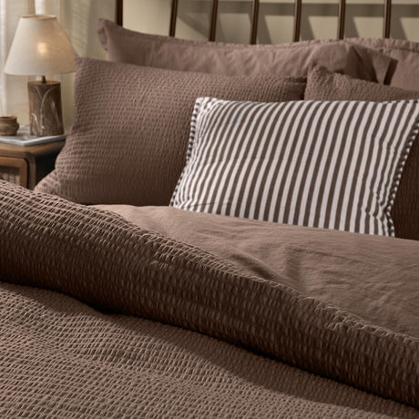 Hebden Cotton Plain Duvet Cover Set in cocoa brown, shown on a bed with textured seersucker pillowcases and a striped accent cushion in a bedroom setting.