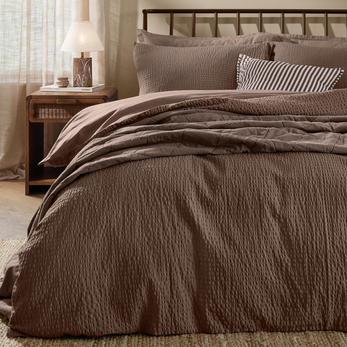 Hebden Cotton Plain Duvet Cover Set in cocoa brown on a bed with striped and textured pillows, dark metal headboard, in a bedroom with wooden nightstand and lamp.