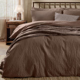 Hebden Cotton Plain Duvet Cover Set in cocoa brown on a bed with striped and textured pillows, dark metal headboard, in a bedroom with wooden nightstand and lamp.