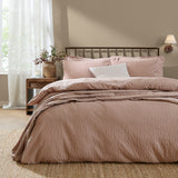 Hebden Cotton Plain Duvet Cover Set in Cotta, a muted pink seersucker-textured cover with matching pillowcases on a bed with dark metal headboard in a bedroom setting.