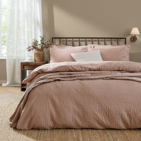 Hebden Cotton Plain Duvet Cover Set in Cotta, a muted pink seersucker-textured cover with matching pillowcases on a bed with dark metal headboard in a bedroom setting.