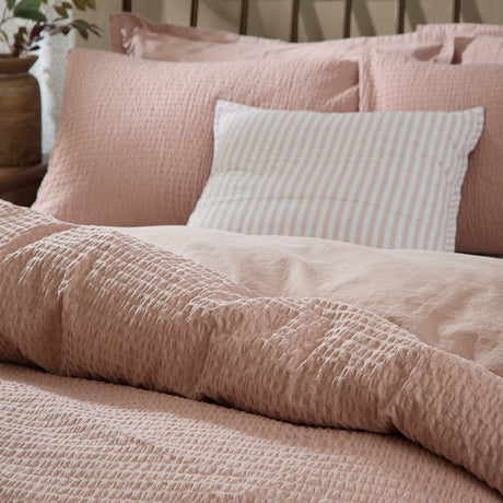 Hebden Cotton Plain Duvet Cover Set in Cotta pink, seersucker-textured cotton with puckered surface, shown on a bed with matching pillowcases and a striped cushion.