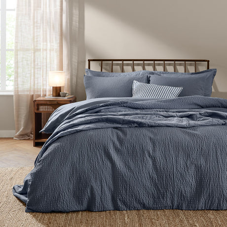 Hebden Cotton Plain Duvet Cover Set in denim blue with seersucker texture, styled on a bed with matching pillowcases and a striped cushion in a bedroom setting.