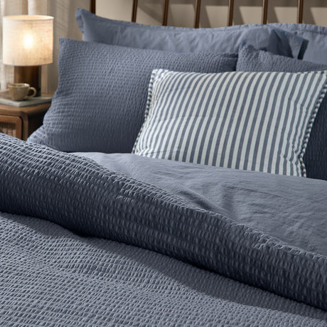 Hebden Cotton Plain Duvet Cover Set in denim blue with textured seersucker finish, shown on a bed with a striped accent cushion in a bedroom setting.