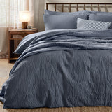 Hebden Cotton Plain Duvet Cover Set in Denim Blue with seersucker texture, dressed on a bed with matching pillowcases and a striped cushion in a bedroom setting.