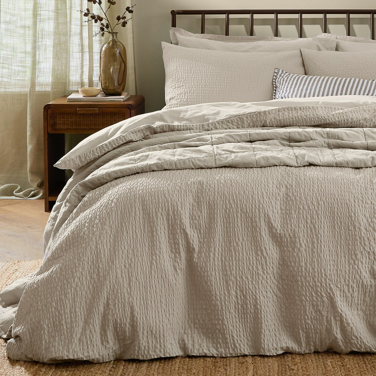 Hebden Cotton Plain Duvet Cover Set in stone on a bed with striped accent cushions, showing the seersucker-textured cotton cover and matching pillowcases in a bedroom setting.
