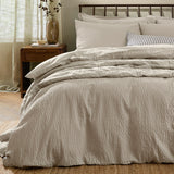 Hebden Cotton Plain Duvet Cover Set in stone on a bed with striped accent cushions, showing the seersucker-textured cotton cover and matching pillowcases in a bedroom setting.