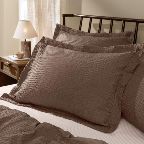 Hebden Percale Cotton Pillowcase Pair in cocoa brown with textured seersucker fronts, shown propped against a dark metal bed frame in a bedroom setting.
