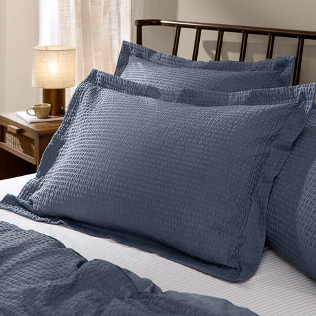 Hebden Percale Cotton Pillowcase Pair in denim blue with a textured seersucker finish, styled on a bed beside a matching duvet in a bedroom setting.