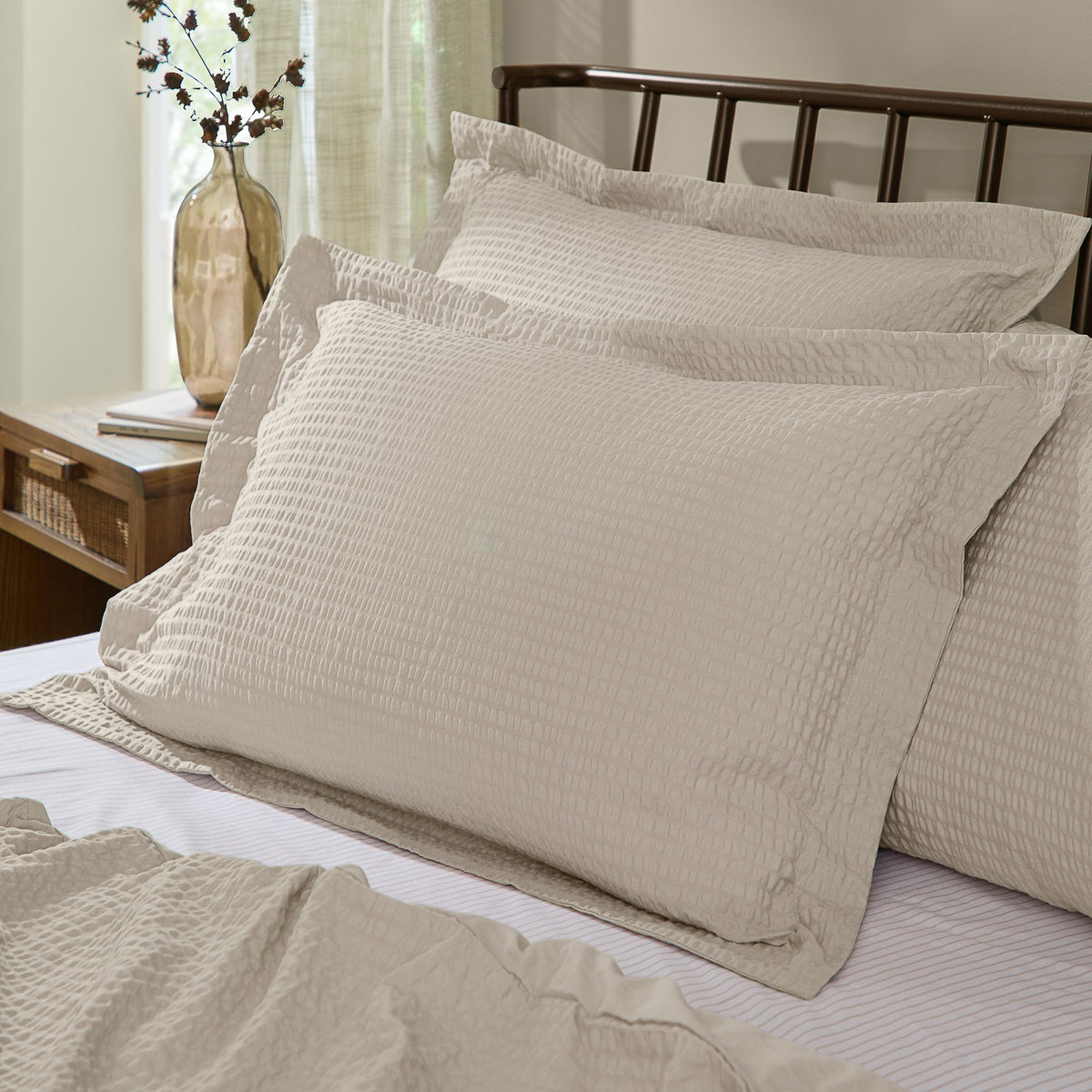 Hebden Percale Cotton Pillowcase Pair in stone, shown with textured seersucker-effect shams styled on a bed with a dark metal headboard in a bedroom setting.