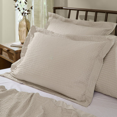 Hebden Percale Cotton Pillowcase Pair in stone, shown with textured seersucker-effect shams styled on a bed with a dark metal headboard in a bedroom setting.