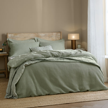 Hebden Cotton Plain Duvet Cover Set in Bay Green sage, waffle-weave cotton bedding with matching pillowcases and striped cushion, styled on a bed with wooden headboard in a bedroom setting.