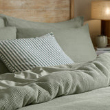 Hebden Cotton Plain Duvet Cover Set in Bay Green, showing waffle-weave sage cotton bedding with striped cushion, plain pillowcases and a warm-lit bedroom setting.