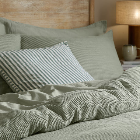Hebden Cotton Plain Duvet Cover Set in Bay Green, showing waffle-weave sage cotton bedding with striped cushion, plain pillowcases and a warm-lit bedroom setting.