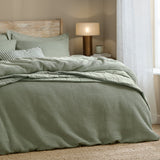 Hebden Cotton Plain Duvet Cover Set in Bay Green, sage waffle-weave cover with striped and plain cushions on a bed in a bedroom with warm lamp lighting.
