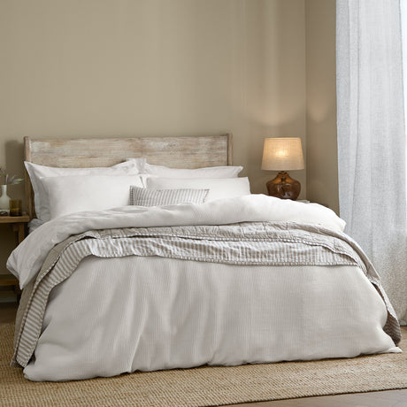 Hebden Cotton Plain Duvet Cover Set in Chalk on a bed with wooden headboard, layered with striped cushions in a neutral bedroom setting.
