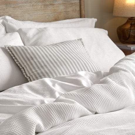 Hebden Cotton Plain Duvet Cover Set in Chalk, shown in a bedroom with waffle-textured white bedding, a striped bolster cushion, and a wooden headboard.