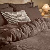 Hebden Cotton Plain Duvet Cover Set in Cocoa Brown, waffle-textured cover with striped accent cushion on a bed, styled in a bedroom with warm lamp lighting.