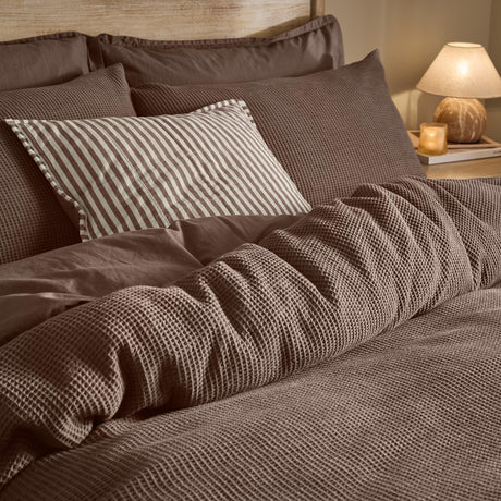 Hebden Cotton Plain Duvet Cover Set in Cocoa Brown, waffle-textured cover with striped accent cushion on a bed, styled in a bedroom with warm lamp lighting.
