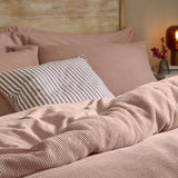Hebden Cotton Plain Duvet Cover Set in Cotta dusky pink, waffle-weave texture shown on a bed with a striped cushion and wooden headboard.