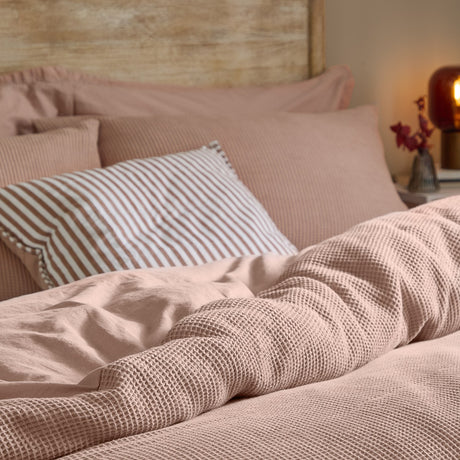 Hebden Cotton Plain Duvet Cover Set in Cotta dusky pink, waffle-weave texture shown on a bed with a striped cushion and wooden headboard.