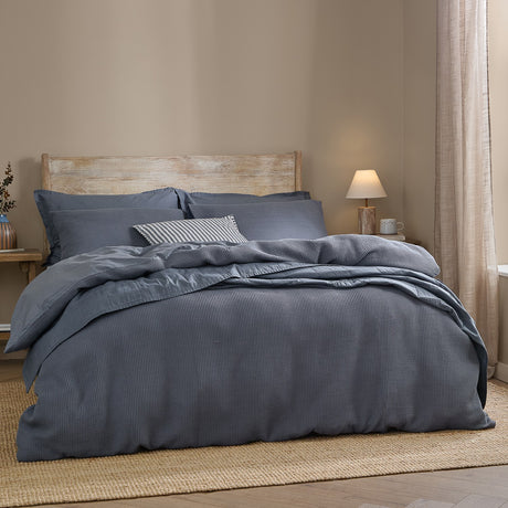 Hebden Cotton Plain Duvet Cover Set in denim blue with waffle-textured face, styled on a bed with matching pillowcases and a striped cushion in a bedroom setting.