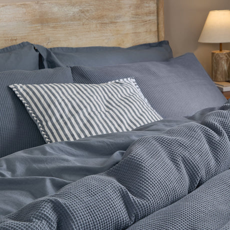 Hebden Cotton Plain Duvet Cover Set in denim blue on a bed, showing the waffle-textured cotton fabric with matching pillowcases and a striped cushion.