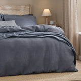 Hebden Cotton Plain Duvet Cover Set in denim blue with waffle-textured face, dressed on a bed beside striped cushions in a bedroom setting.
