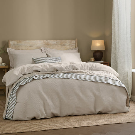 Hebden Cotton Plain Duvet Cover Set in Stone dressing a bed with waffle-textured pillowcases and a striped accent cushion in a neutral bedroom.
