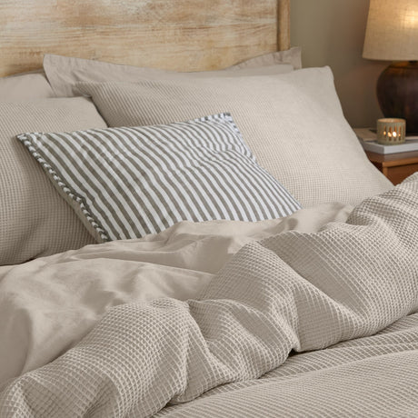Hebden Cotton Plain Duvet Cover Set in Stone, waffle-textured cotton bedding with a striped cushion, styled on a bed in a bedroom setting.
