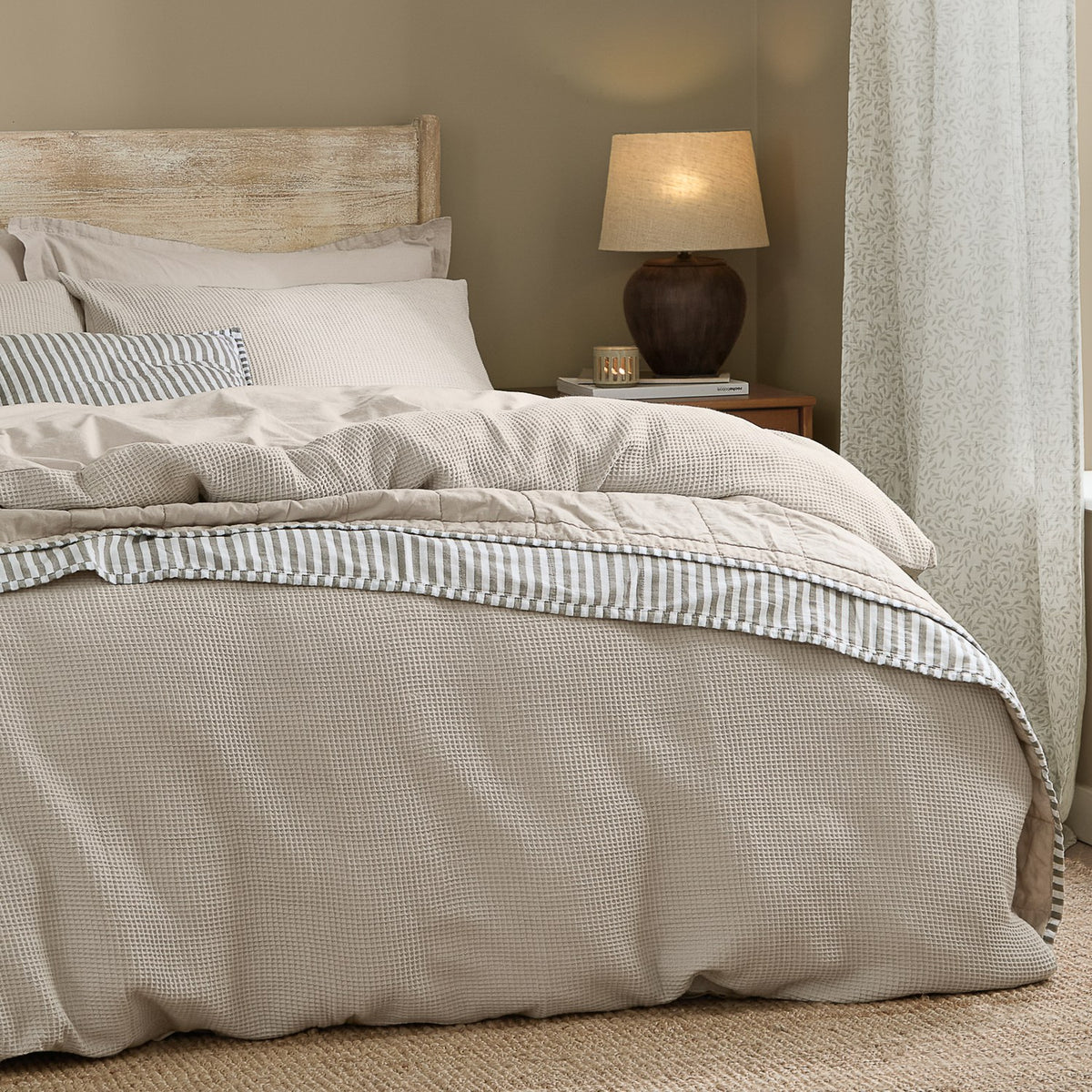 Hebden Cotton Plain Duvet Cover Set in Stone, waffle-textured beige bedding on a bed with striped accent pillow, wooden headboard, and bedside lamp in a neutral bedroom setting.