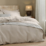Hebden Cotton Plain Duvet Cover Set in Stone, waffle-textured beige bedding on a bed with striped accent pillow, wooden headboard, and bedside lamp in a neutral bedroom setting.