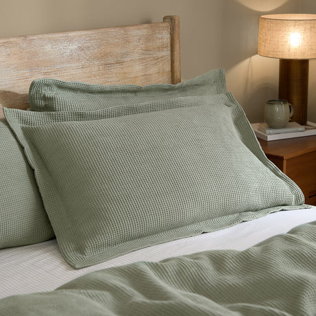 Hebden Percale Cotton Pillowcase Pair in Bay Green shown on a bed with waffle-textured cotton pillowcases against a wooden headboard and bedside lamp.