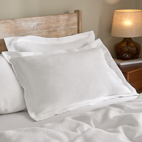 Hebden Percale Cotton Pillowcase Pair in Chalk with waffle-textured front, shown styled on a bed with white bedding in a bedroom setting with wooden headboard.