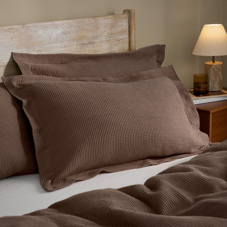 Hebden Percale Cotton Pillowcase Pair in cocoa brown with waffle-textured face, styled on a bed with a matching waffle throw beside a wooden headboard and bedside lamp.