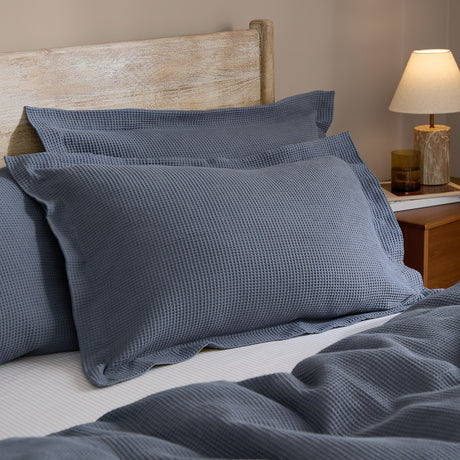 Hebden Percale Cotton Pillowcase Pair in denim blue, shown with waffle-weave texture on a bed against a wooden headboard and bedside lamp.