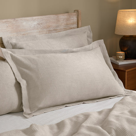 Hebden waffle-textured cotton pillowcase pair in stone, styled on a bed with a wood headboard, oxford-style flanged edges, beside a bedside lamp.