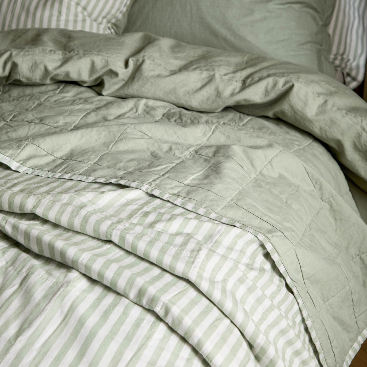 Hebden Percale Cotton Bedspread in eucalyptus/bay green, shown reversible with a quilted plain green face folded back to reveal the pale green and white striped underside on a bed.