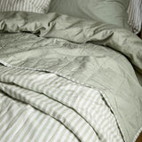 Hebden Percale Cotton Bedspread in eucalyptus/bay green, shown reversible with a quilted plain green face folded back to reveal the pale green and white striped underside on a bed.