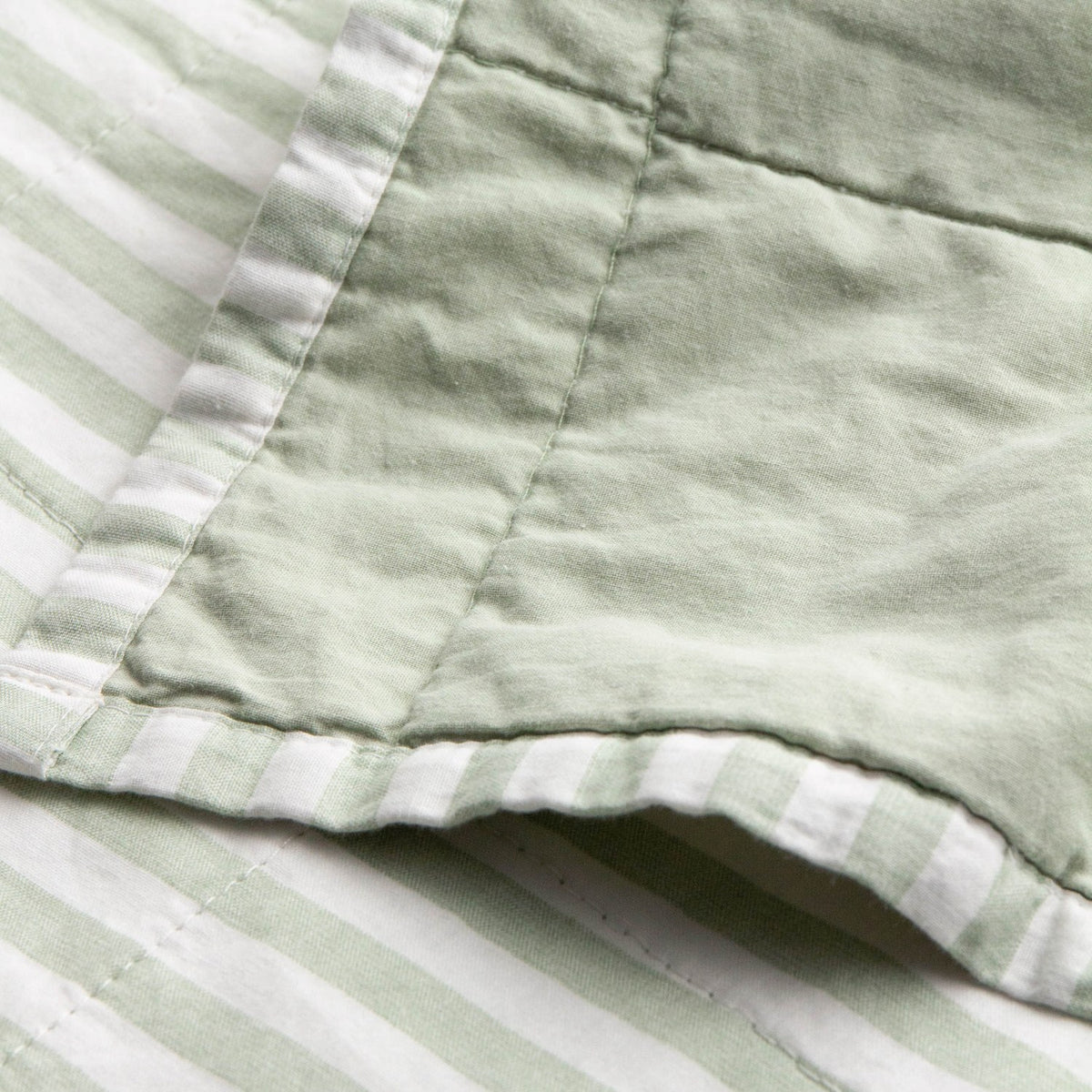 Close-up of the Hebden Percale Cotton Bedspread in Eucalyptus/Bay Green, showing quilted stitching where the plain green face meets the reversible green-and-white striped edge.