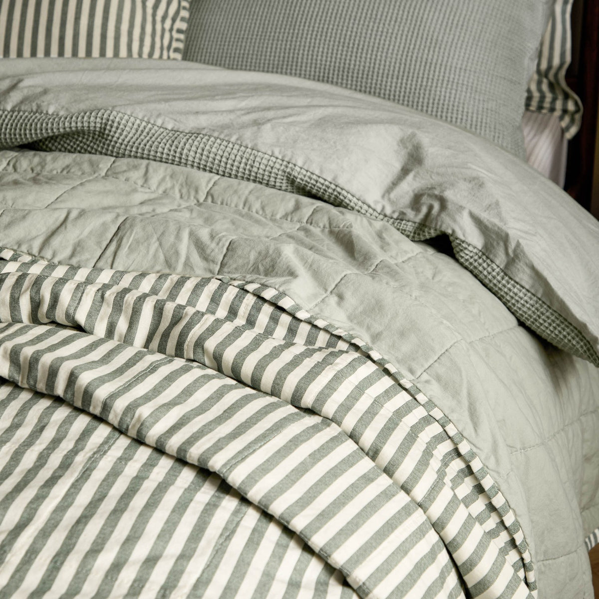 Hebden Percale Cotton Bedspread in Forest/Bay Green, shown folded on a bed with striped cotton reverse and quilted plain green face, paired with gingham-check pillows.