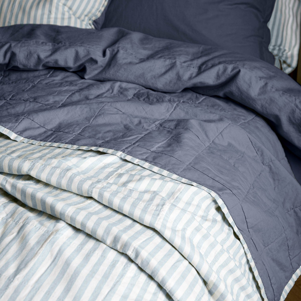 Hebden Percale Cotton Bedspread in denim blue, quilted percale cotton reversing to a pale mineral-blue and white stripe, shown folded back on a bed.