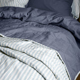Hebden Percale Cotton Bedspread in denim blue, quilted percale cotton reversing to a pale mineral-blue and white stripe, shown folded back on a bed.