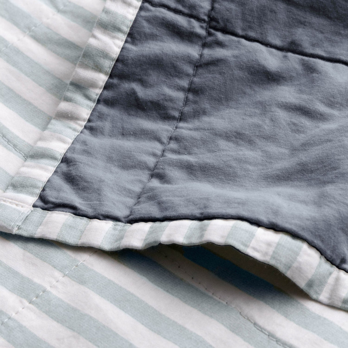 Close-up of the reversible Hebden Percale Cotton Bedspread, showing the denim-blue cotton face folded back over the pale mineral-blue and white striped side with visible stitching.